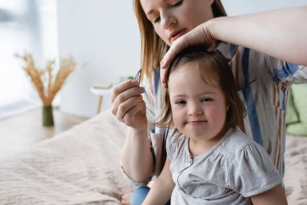 Размытая мать держит заколку для волос рядом с дочерью с синдромом Дауна в спальне — стоковое фото