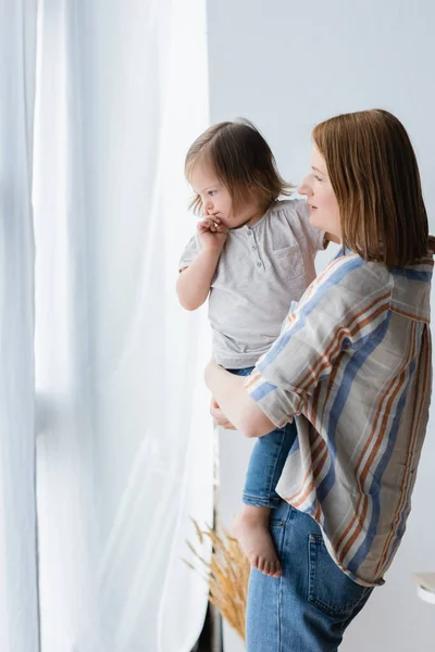 Mujer sosteniendo hija con síndrome de Down cerca de cortinas en casa - foto de stock