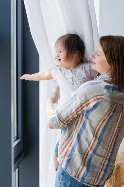 Woman holding child with down syndrome near window at home — Stock Photo