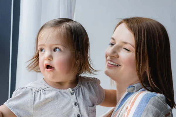 Весела жінка на розмитому тлі дивиться на дочку з синдромом Дауна вдома — стокове фото
