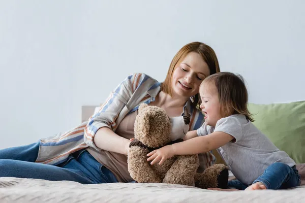 Mulher sorridente brincando com a filha com síndrome de down e brinquedo macio na cama — Fotografia de Stock