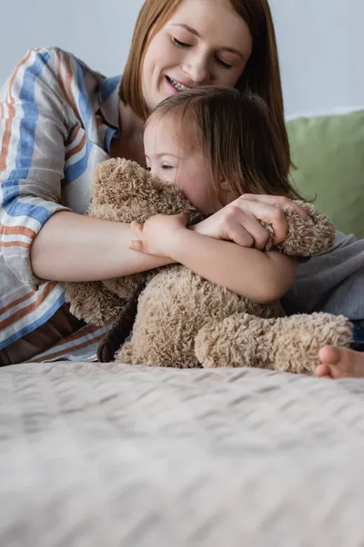 Улыбающаяся женщина обнимает дочь с синдромом Дауна и мягкой игрушкой на кровати — стоковое фото