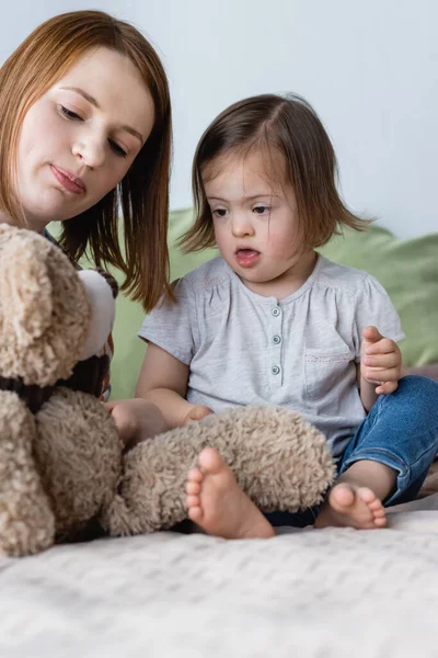 Родитель держит плюшевого мишку рядом с ребенком с синдромом Дауна на кровати — стоковое фото