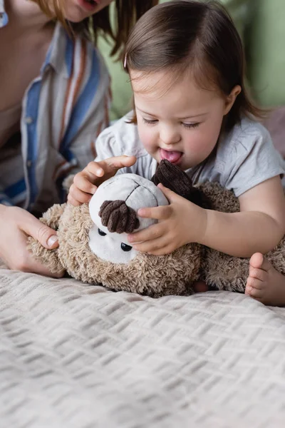 Menina do bebê com síndrome de down segurando brinquedo macio perto mãe turva na cama — Fotografia de Stock