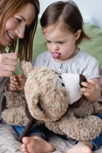 Улыбающаяся женщина играет с маленькой дочерью с синдромом Дауна и мягкой игрушкой на кровати — стоковое фото