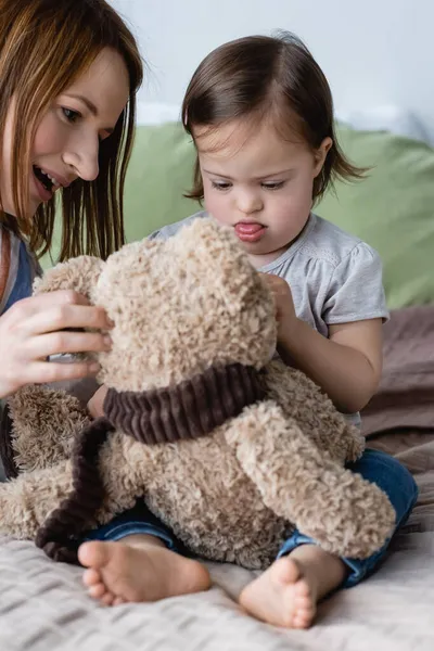 Girl with down syndrome playing with soft toy near mother on bed — Stock Photo