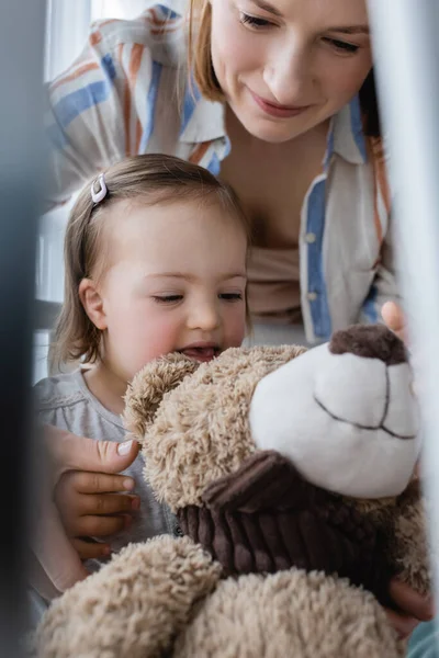 Fröhliches Kind mit Down-Syndrom schaut Teddybär bei Mutter zu Hause an — Stockfoto