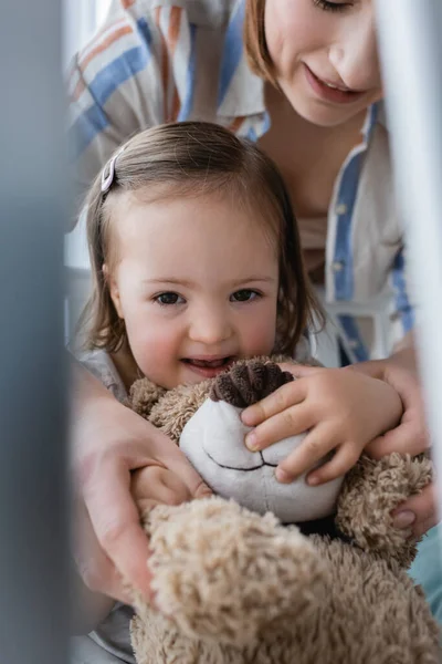 Positive child with down syndrome holding soft toy near smiling mother in baby crib — Stock Photo