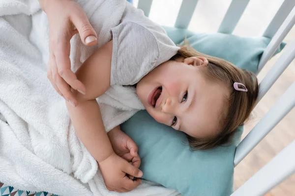 Вид сверху положительного ребенка с синдромом пуха, лежащего на кроватке рядом с расплывчатой мамой — стоковое фото