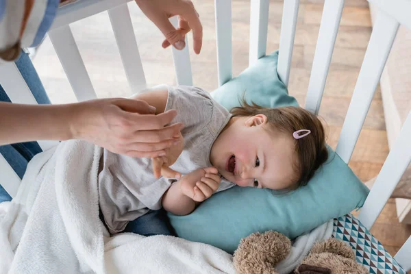 Top view of mother holding hand of cheerful child with down syndrome in baby crib — Stock Photo