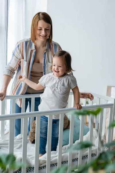 Улыбающаяся женщина стоит рядом с дочерью с синдромом Дауна в детской кроватке — стоковое фото