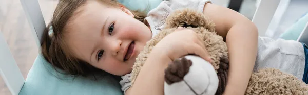 Вид зверху на дівчину з синдромом Дауна, що обіймає м'яку іграшку в ліжечку, банер — стокове фото