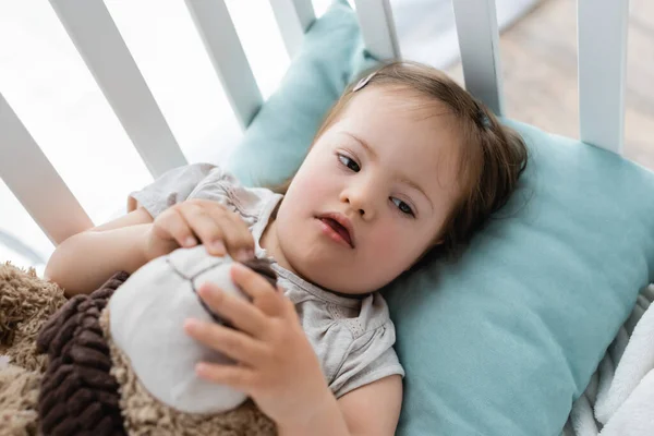 Високий кут зору дитини з синдромом Дауна, що тримає розмиту м'яку іграшку в дитячому ліжку — стокове фото