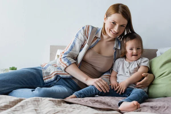 Lächelnde Mutter blickt in die Kamera, während sie Tochter mit Down-Syndrom auf dem Bett umarmt — Stockfoto
