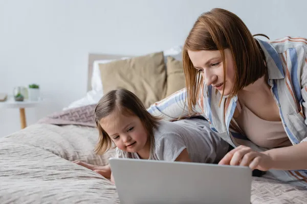 Freelancer usando laptop cerca de hija con síndrome de Down en la cama - foto de stock