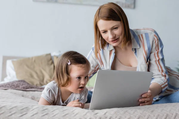 Baby mit Down-Syndrom blickt im Schlafzimmer auf Laptop neben verschwommener Mutter — Stockfoto