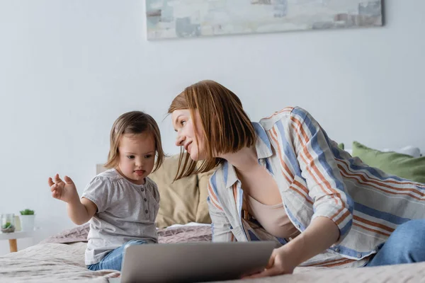 Mujer sonriente mirando a un niño con síndrome de Down cerca de la computadora portátil borrosa en el dormitorio - foto de stock