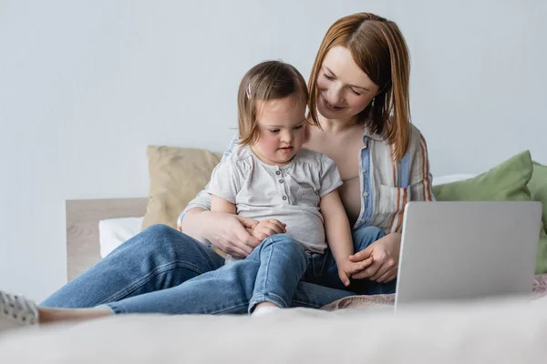 Femme positive regardant l'enfant avec le syndrome du duvet près de l'ordinateur portable sur le lit — Photo de stock