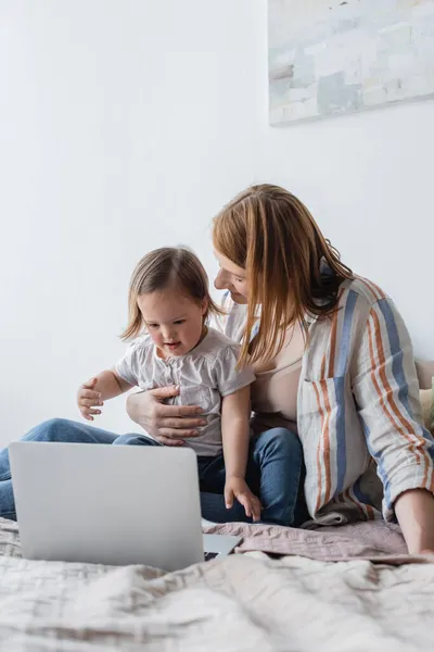 Lächelnde Frau umarmt Kleinkind mit Down-Syndrom in der Nähe von Laptop auf verschwommenem Bett — Stockfoto