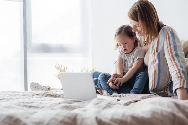 Padres felices apuntando al portátil cerca de la hija con síndrome de Down en la cama - foto de stock