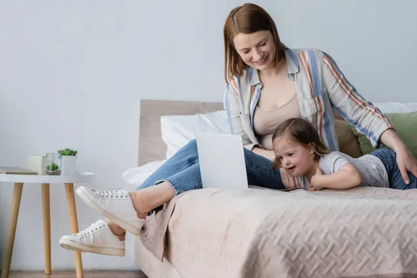 Родитель улыбается рядом с дочерью с синдромом Дауна и ноутбуком на кровати — стоковое фото