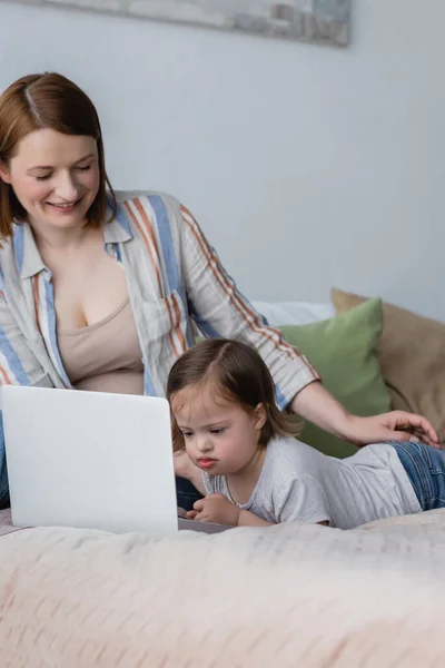 Child with down syndrome looking at laptop near positive parent in bedroom — Stock Photo
