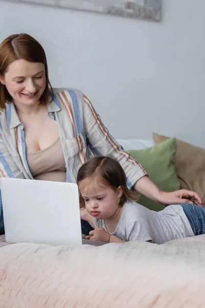 Niño con síndrome de Down mirando portátil cerca de padres positivos en el dormitorio - foto de stock