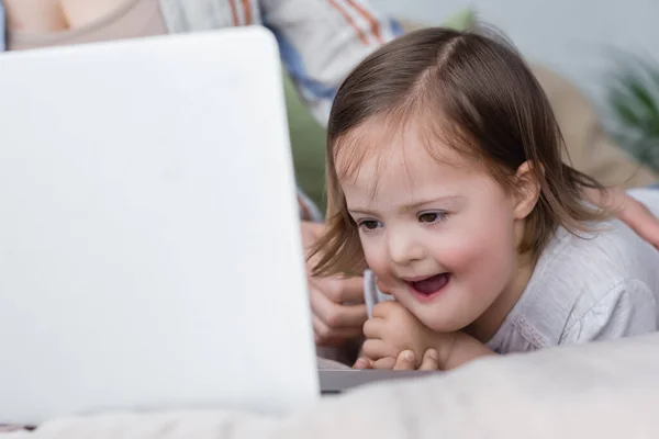 Aufgeregtes Kind mit Down-Syndrom blickt nahe Mutter auf verschwommenen Laptop — Stockfoto