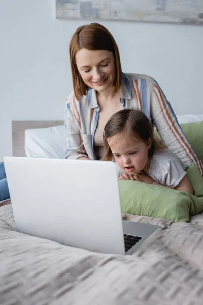 Freelancer pai sorridente olhando para a filha com síndrome de down perto laptop na cama — Fotografia de Stock