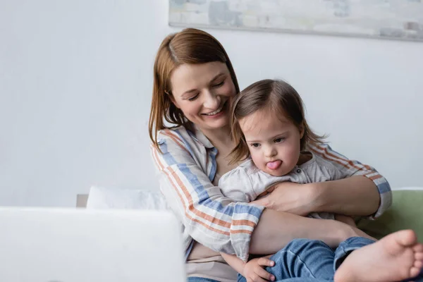 Smiling mother hugging daughter with down syndrome sticking out tongue near blurred laptop — Stock Photo