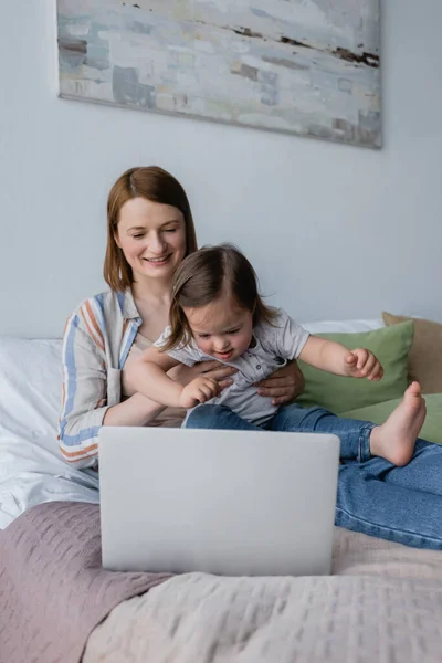 Lächelnde Frau hält Kind mit Down-Syndrom neben Laptop im Schlafzimmer — Stockfoto