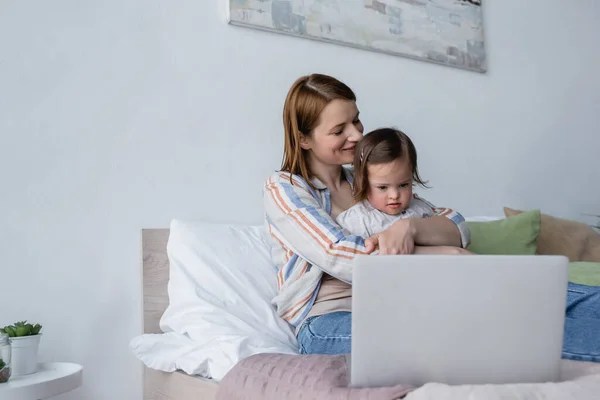 Щаслива мама обіймає дочку малюка біля ноутбука на ліжку — стокове фото