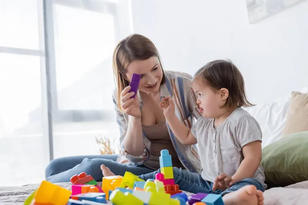 Sonriente madre sosteniendo bloque de construcción cerca de un niño con síndrome de Down en la cama - foto de stock