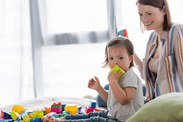 Niño con síndrome de Down jugando bloques de construcción cerca de la madre sonriente en la cama - foto de stock