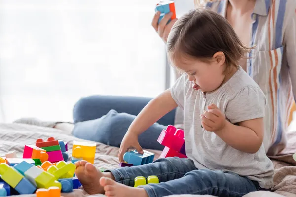 Child with down syndrome sticking out tongue while playing building blocks near mother on bed — Stock Photo