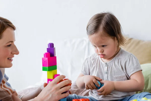 Niño enfocado con síndrome de Down mirando bloques de construcción cerca de la mamá sonriente en el dormitorio - foto de stock