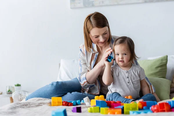 Smiling parent holding building block near daughter with down syndrome on bed — Stock Photo