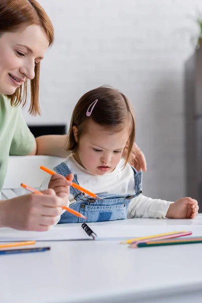 Mujer sonriente sosteniendo lápiz de color cerca de su hija con síndrome de Down y papel en casa - foto de stock