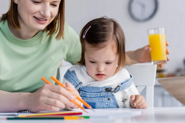 Enfant avec le syndrome du duvet dessin sur papier près de maman souriante avec du jus d'orange dans la cuisine — Photo de stock