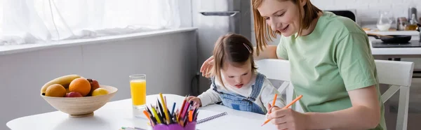 Padres e hijos con síndrome de Down dibujando en papel cerca del jugo de naranja y frutas en la cocina, pancarta - foto de stock