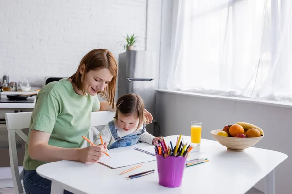 Niño con síndrome de Down dibujo sobre papel cerca de la madre y frutas en la cocina - foto de stock