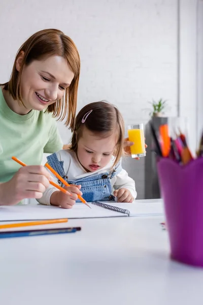 Happy woman with orange juice drawing near daughter with down syndrome in kitchen — Stock Photo