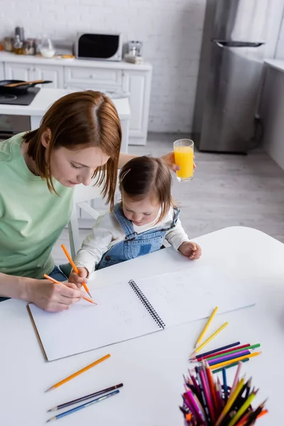 High angle view of girl with down syndrome drawing near mom with orange juice in kitchen — Stock Photo