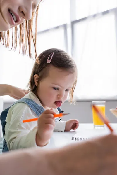 Criança com síndrome de down segurando lápis de cor perto da mãe e suco de laranja borrado — Fotografia de Stock