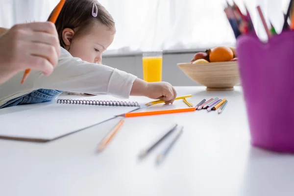 Девушка с синдромом Дауна берет цветной карандаш рядом с фруктами, апельсиновый сок и родителя дома — стоковое фото