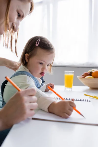 Criança com síndrome de down desenho em papel perto de mãe e suco de laranja na cozinha — Fotografia de Stock