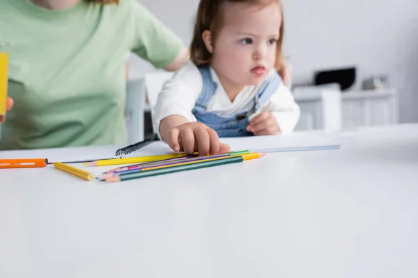 Цветные карандаши рядом с размытым ребенком с синдромом Дауна и родителем на кухне — стоковое фото