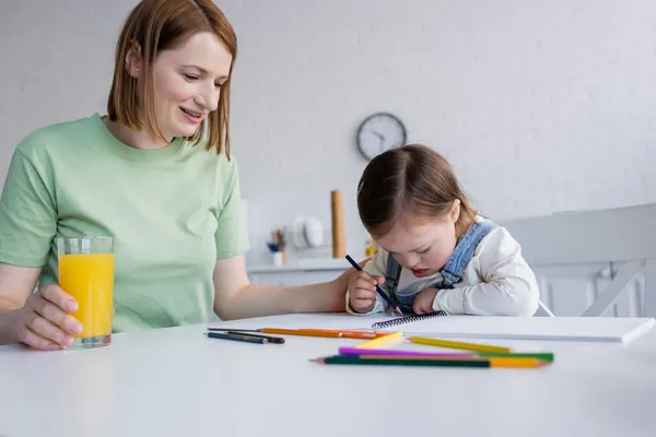 Donna positiva che tiene il vetro di succo d'arancia vicino a figlia con sindrome giù che disegna in cucina — Foto stock