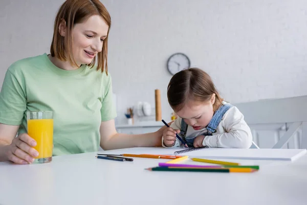 Mujer positiva sosteniendo vaso de jugo de naranja cerca de hija con síndrome de Down dibujo en la cocina - foto de stock