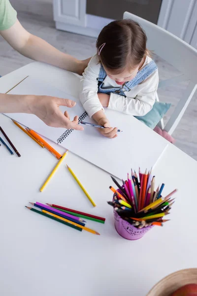 Вид зверху дитини з синдромом вниз, що малює на папері біля мами вдома — стокове фото